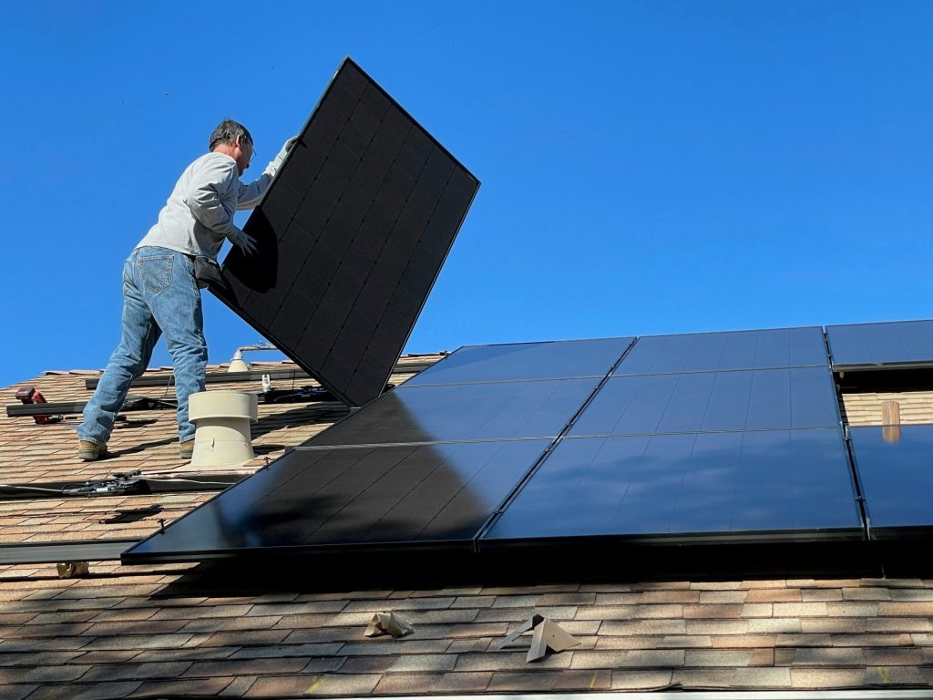 A person installing a black solar panel on a shingled roof under blue sky